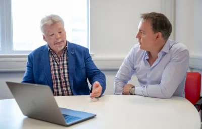 SImon Weston CBE in mid blue jacket with laptop in meeting with man in light blue shirt perched on a red char in an office.