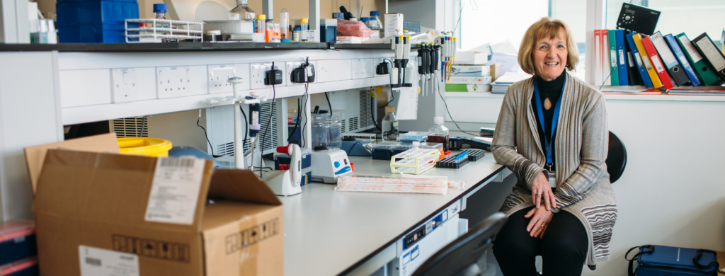 Professor Janet Lord wears a black top and grey cardigan as she sits at a desk covered in research equipment. Janet smiles at the camera.