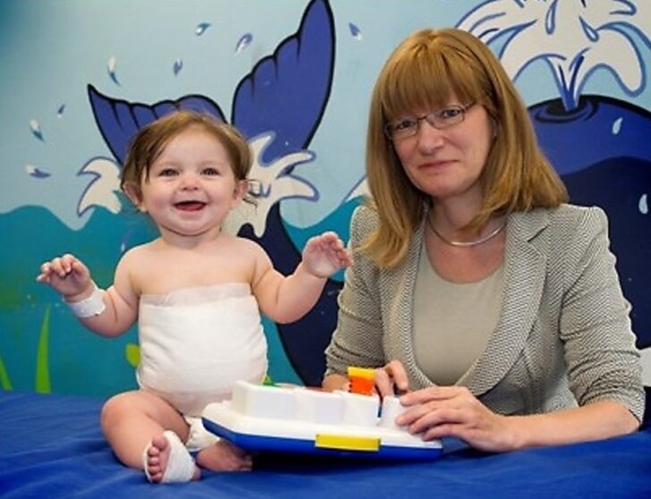 Professor Amber Young sits at a blue table, wearing a neutral blazer suit, in front of a wall painted with a blue whale. On the table is a smiling baby, who had bandages round its middle.