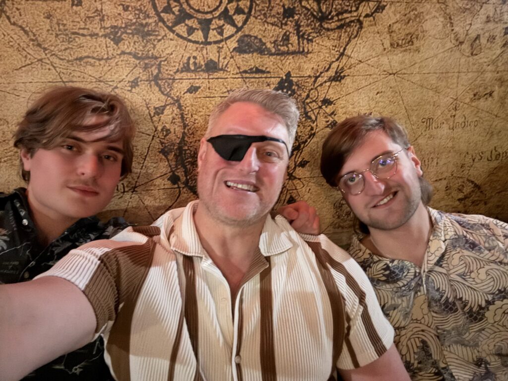 Dan Jackson sits in between his two sons and takes a selfie. There is an old-style map on the wall behind them. Dan is proudly wearing his black eye patch.