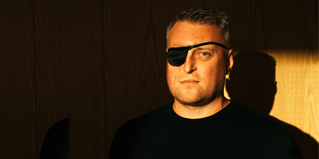 Ambassador Dan Jackson stands in sunlight in front of a wood panelled wall. Dan wears a black t-shirt and a black eye patch.