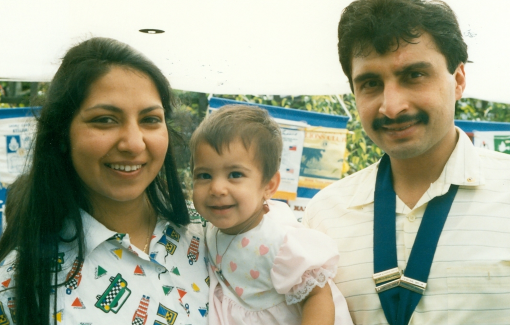 Hemani's mum and dad hold baby Hemani between them. They all smile at the camera.