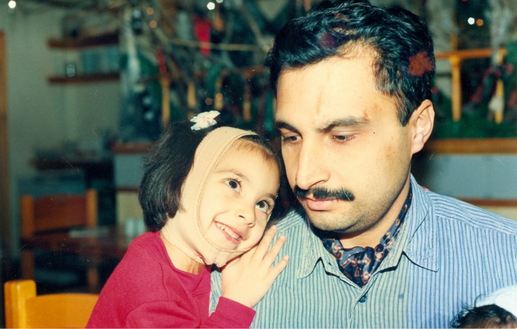 Toddler Hemani leans on her Dad's shoulder and looks up at him smiling. She wears a red jumper, and a bandage wrapped around her head covering her injured ear. Her dad wears a blue jumper.