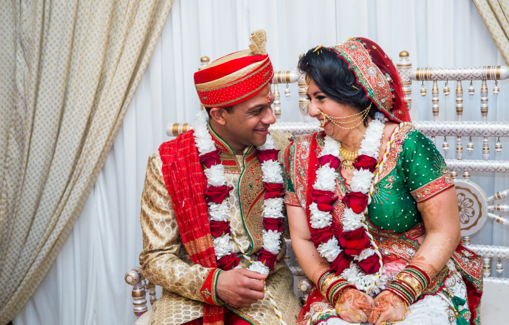 Hemani and her husband wear bright, colourful clothes for their wedding day. They are seated, smiling and laughing with each other.