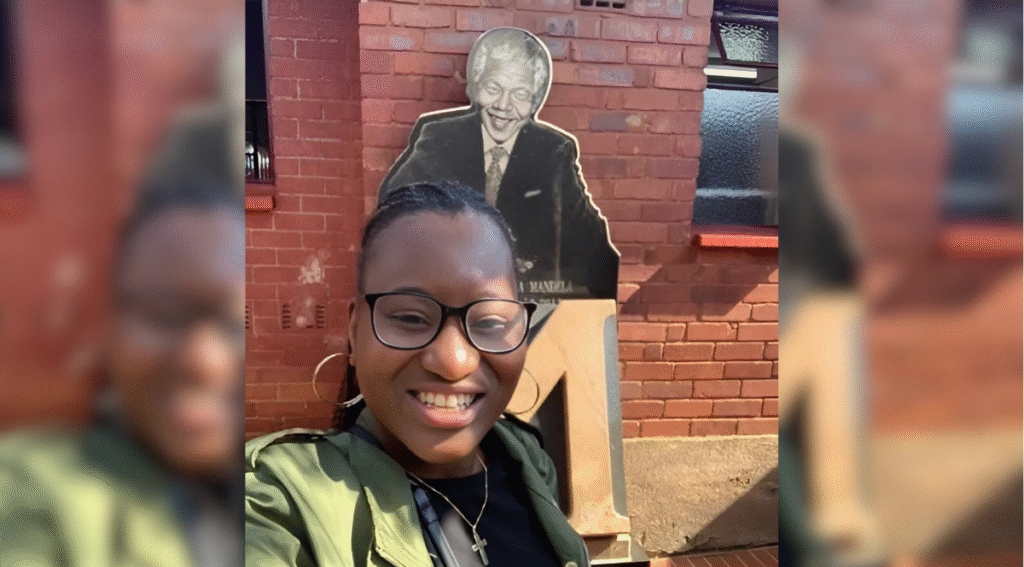 Marian Adejokun smiles at the camera wearing a green coat, black top, as she stands in front of a cutout of Nelson Mandela