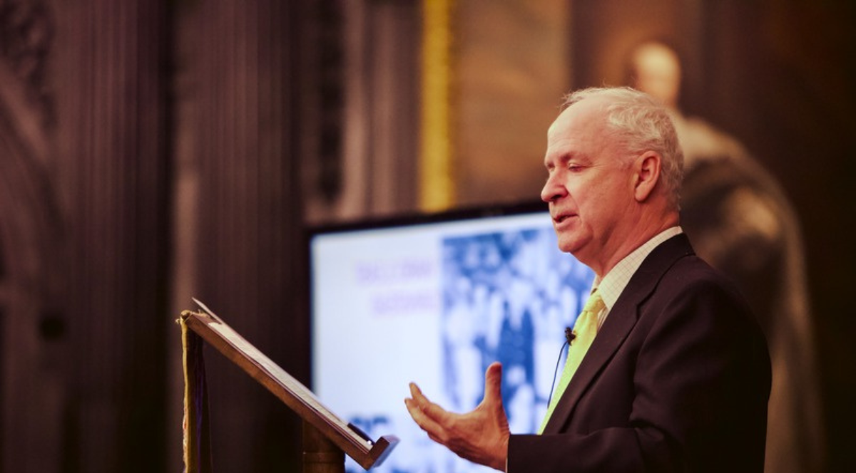 Simon Wessley stands in front of a lecturn to deliver a lecture. A screen with his slides on is in the background.