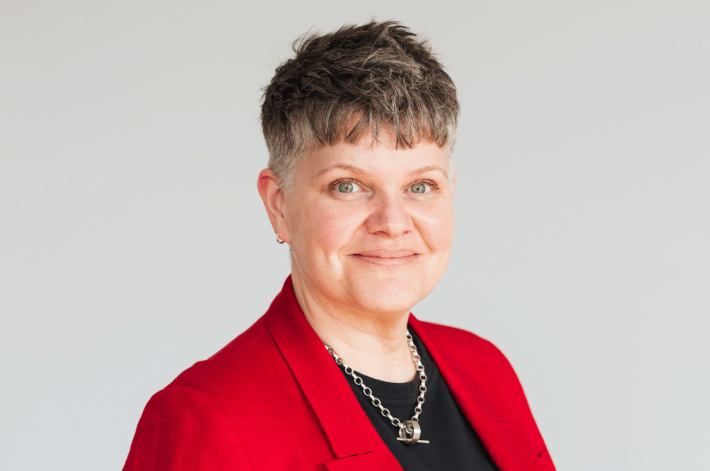 Charlotte Coates wears a red jacket, black top, silver necklace with a hoop and smiles at the camera. Charlotte is the Deputy Chief Executive of The Scar Free Foundation
