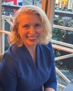Bridget Edminson, Trustee of The Scar Free Foundation, smiles at the camera. Bridget is seated on a balcony overlooking a city street below and wears a blue dress. 