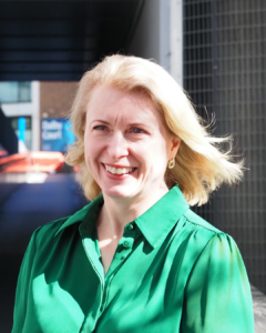 Bridget Edminson, Trustee of The Scar Free Foundation, stands outside grey university buildings in a breeze. She wears a bright green shirt and smiles at the camera. 