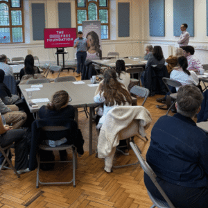Mr Simon Filson stands at the front of the room next to a red screen with white text reading 'The scar free foundation'. Attendees are seated and listen. Simon wears navy blue trousers and a light blue shirt.