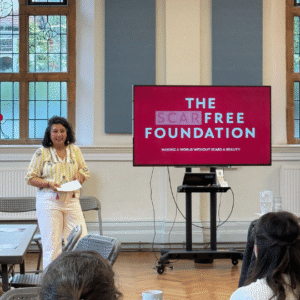Dr Hemani Modasia-Shah speaks to the room standing beside a red screen with 'The Scar Free Foundation' written on it in white text. Hemani is a GP, scar free foundation ambassador and trustee. she wears white trousers and a yellow blouse.