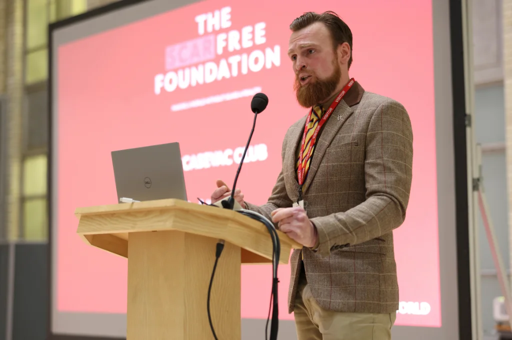 Man speaking at podium with laptop during presentation, large pink screen behind displaying "The Free Foundation" text.