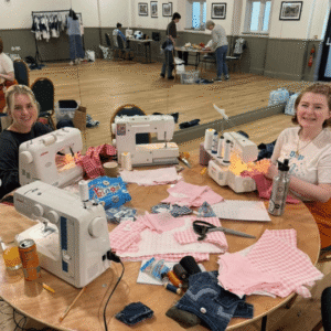 Two volunteers sit in a wood panelled room at a wooden table covered in fabric and sewing machines