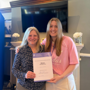 Hannah smiles at the camera and she proudly holds her prize along with another CFSGBI member. Hannah wears a pink t-shirt and white trousers. The CFSGBI member wears a blue patterned blouse and smiles at the camera too.