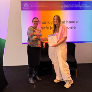 Hannah Waterhouse stands on a stage in front of a multicoloured projection screen and accepts her award from a member of the Craniofacial Society of Great Britain and Ireland. Hannah has long blonde hair, smiles at the camera, and wears a pink Adidas originals t-shirt, white trousers, and white new balance trainers