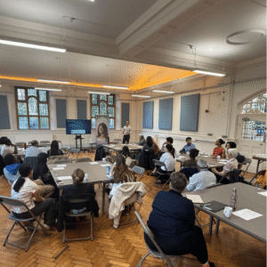 A room full of people sit at chairs around square grey tables to listen to a clinician giving a presentation, who stands at the front beside a screen.