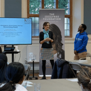 Prof Tanya Shaw and PhD candidate Takunda Ngwenya speak to attendees at the event. They have their 'researching keloids' presentation on screen. Tanya wears black tights, a check skirt, a black jumper and a turquoise scarf, and Takunda wears beige trousers and a bright blue hoodie