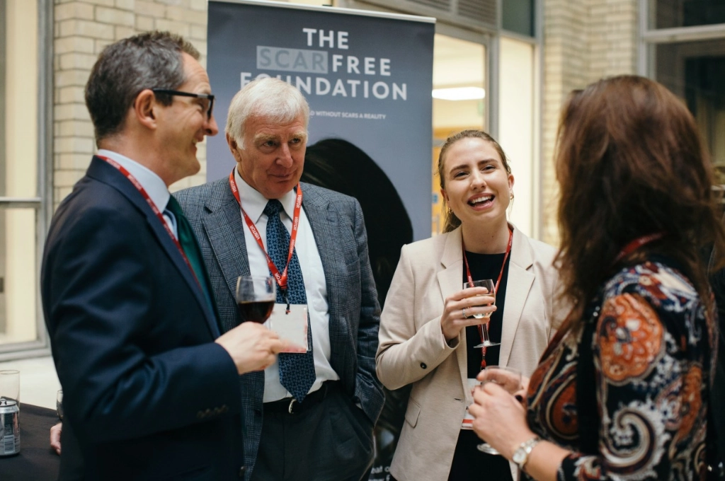 Four professionals networking at a Scar Free Foundation event, holding drinks and conversing indoors.