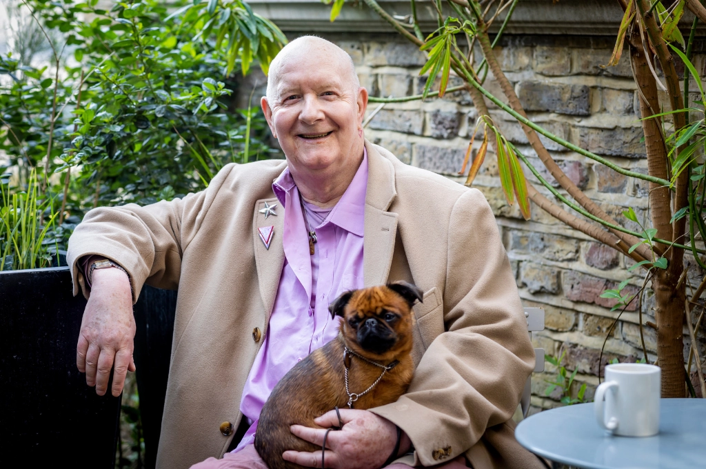Smiling older man in beige blazer and purple shirt holding a brown dog in a garden setting.