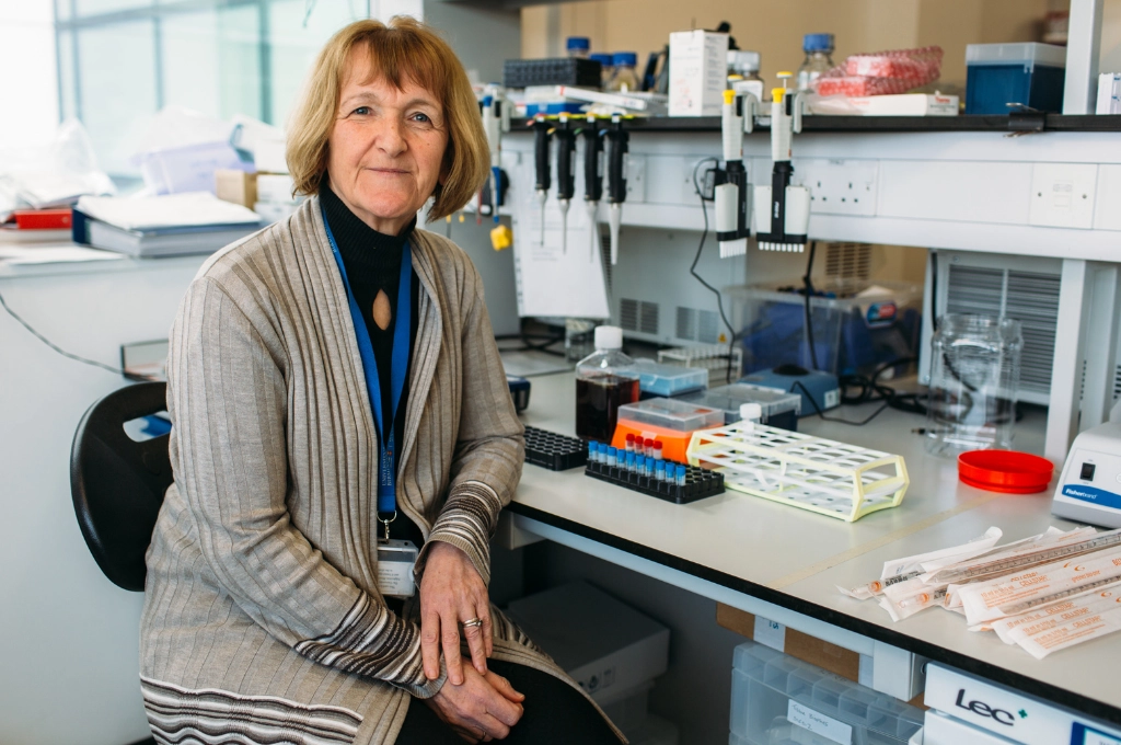 Woman in lab coat at laboratory workstation with scientific equipment and samples, wearing blue ID badge.