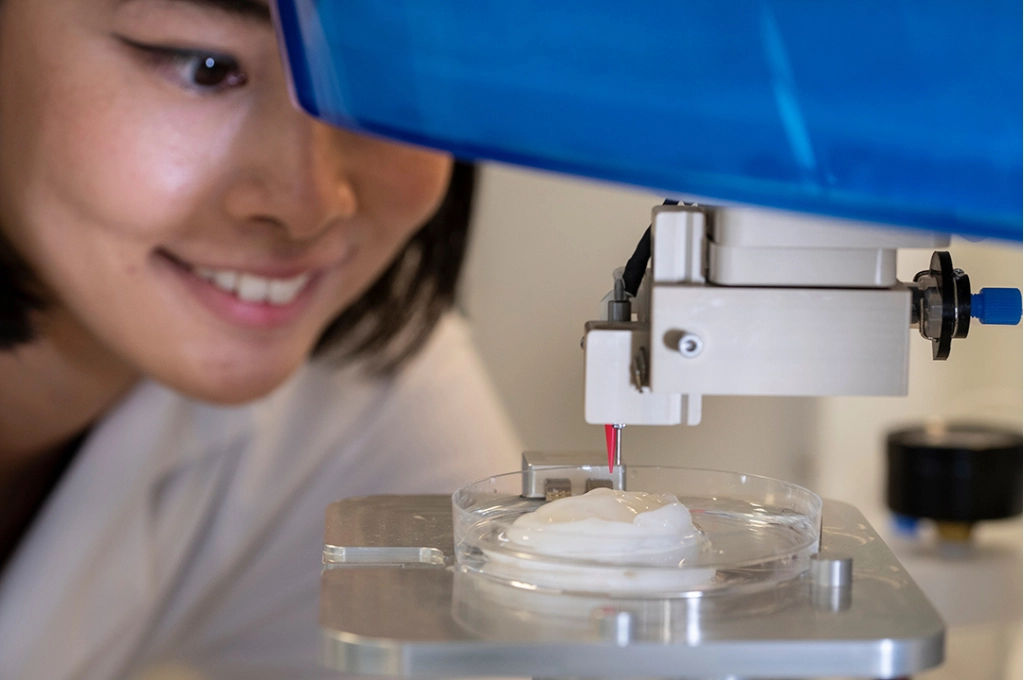 Young woman smiling while observing 3D bioprinter depositing white material onto a petri dish in laboratory setting.