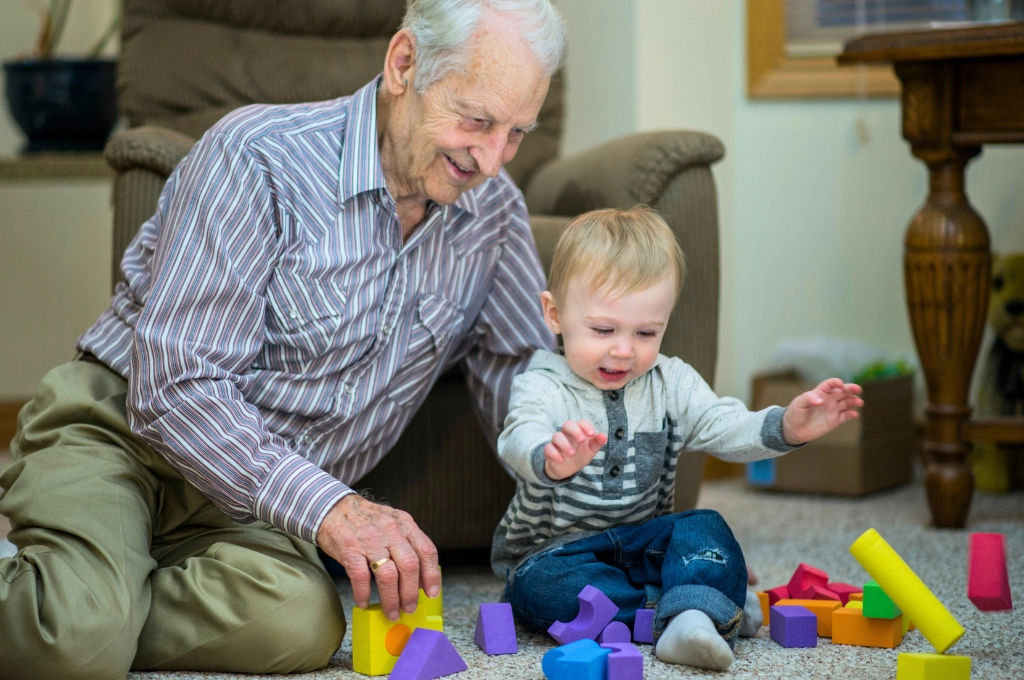 Elderly man and toddler playing with colourful building blocks on living room floor together.