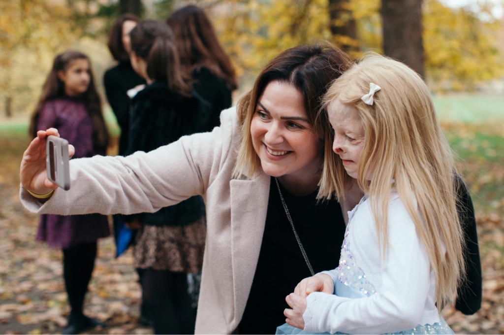 Adult and child taking a selfie together outdoors in autumn setting with other people in background.