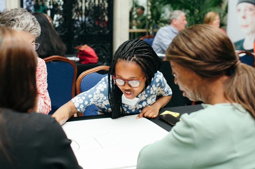 Child with braids and glasses engaged in discussion with adults around a table during a research participation event.