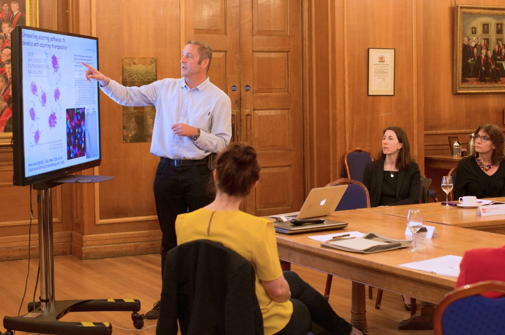 Man presenting microscopy research findings on display screen to seated colleagues in formal meeting room.