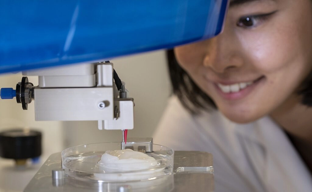 Researcher smiling at laboratory equipment dispensing liquid onto a sample in a petri dish.