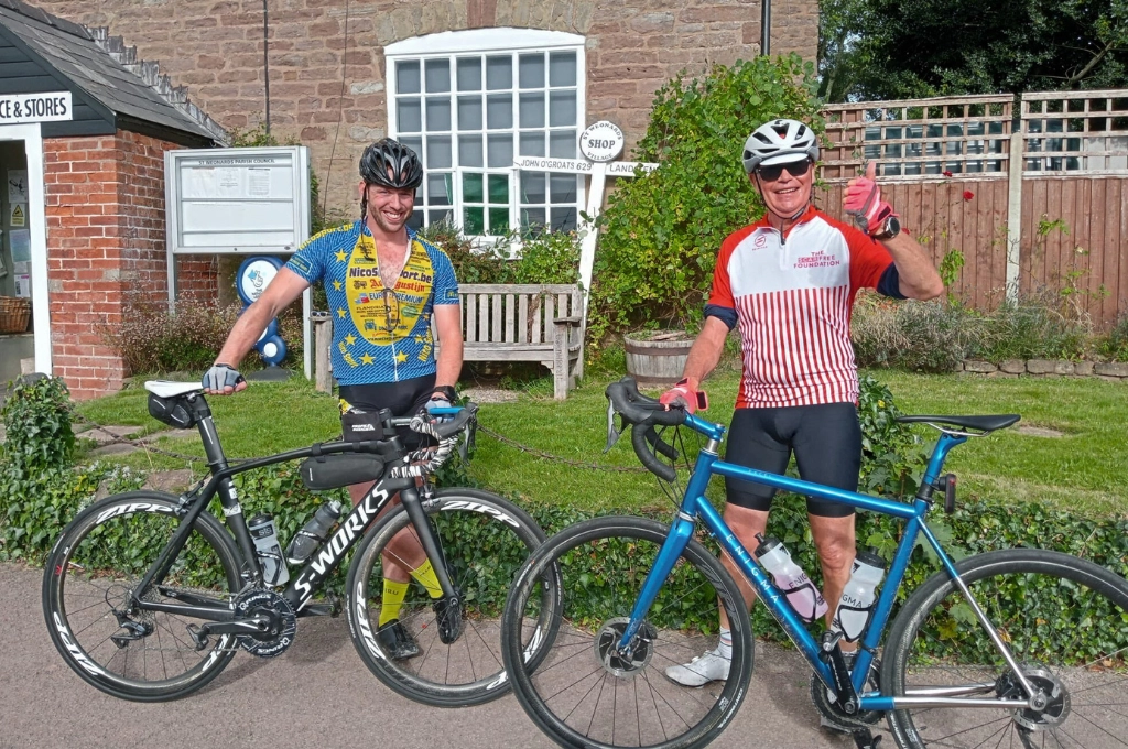 Two cyclists in sports gear with helmets stand with their bikes in a garden setting, ready for a charity ride.
