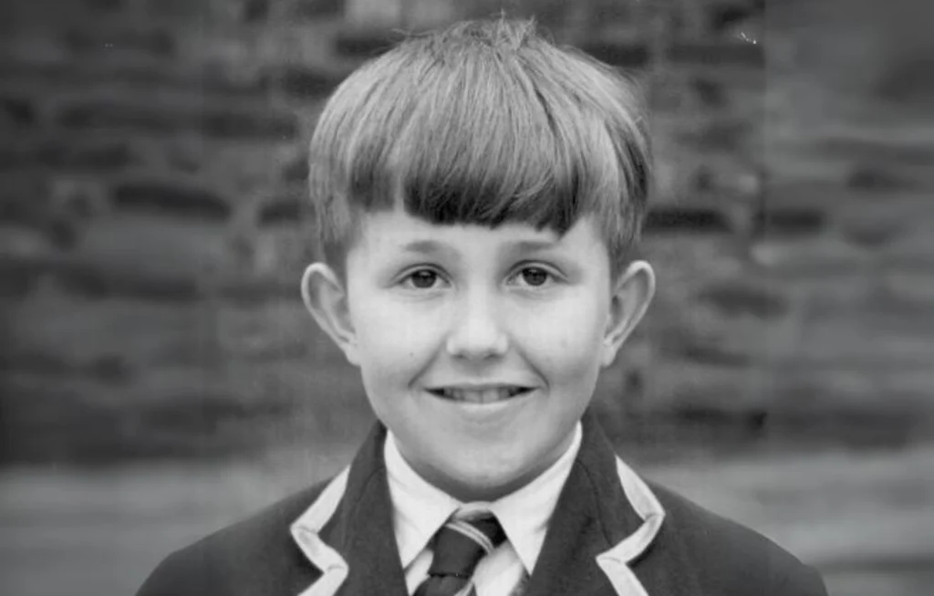 Young boy in school uniform with dark blazer and striped tie, smiling at camera against brick background.