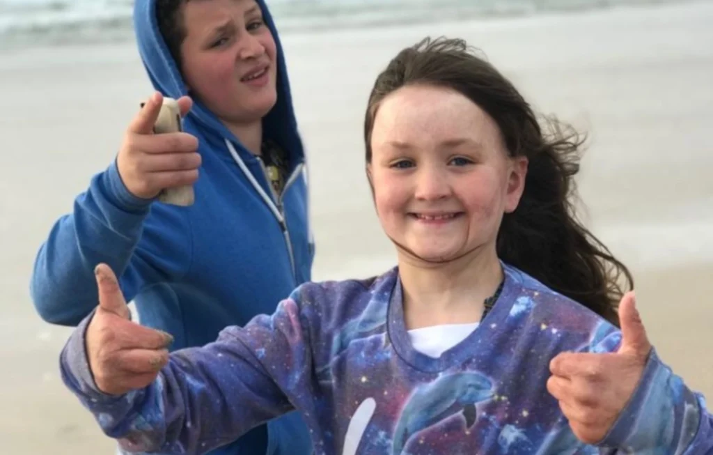 Two young girls at a beach giving thumbs up gestures, both smiling at the camera.