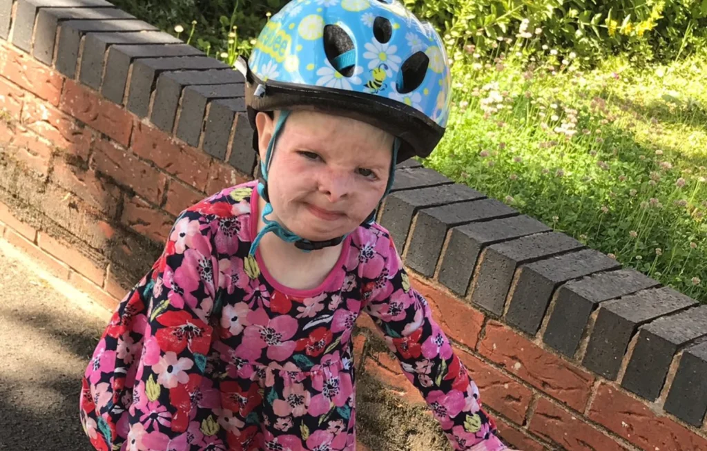 Young child in pink floral shirt and blue bike helmet smiling at camera against brick wall and grass.