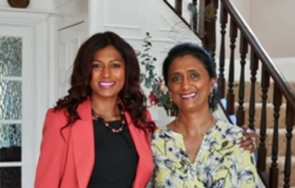 Two women smiling at camera in home setting, one wearing coral blazer, one in yellow patterned top.