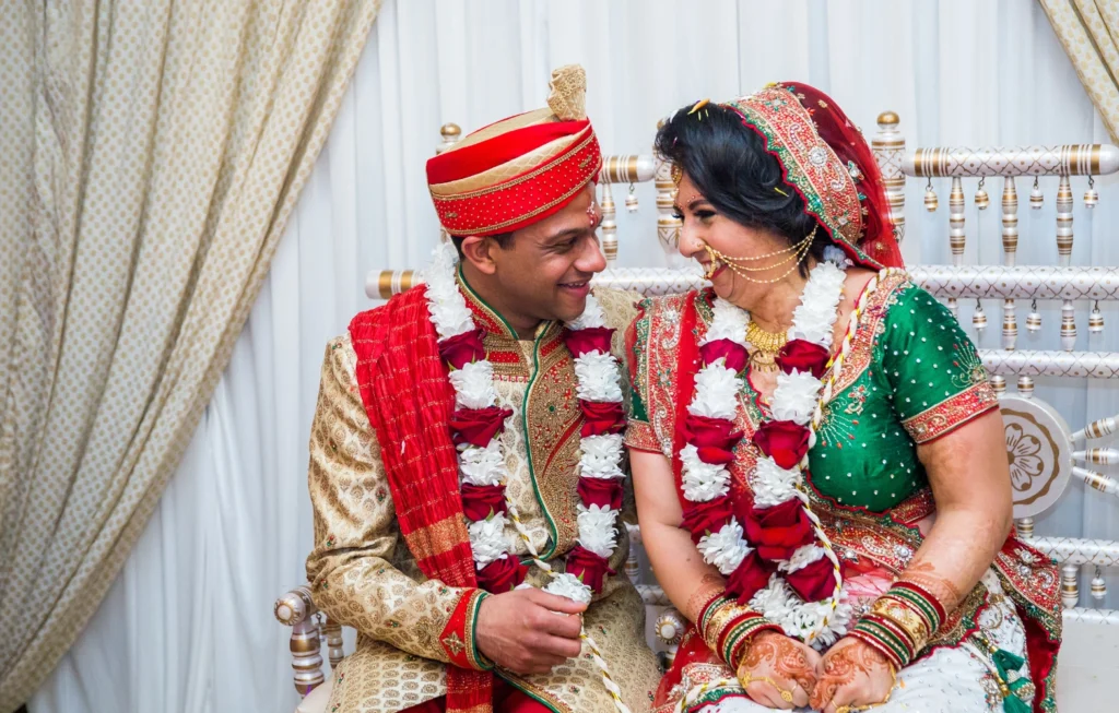 Bride and groom in traditional Indian wedding attire smiling at each other, wearing red and gold garments with flower garlands.