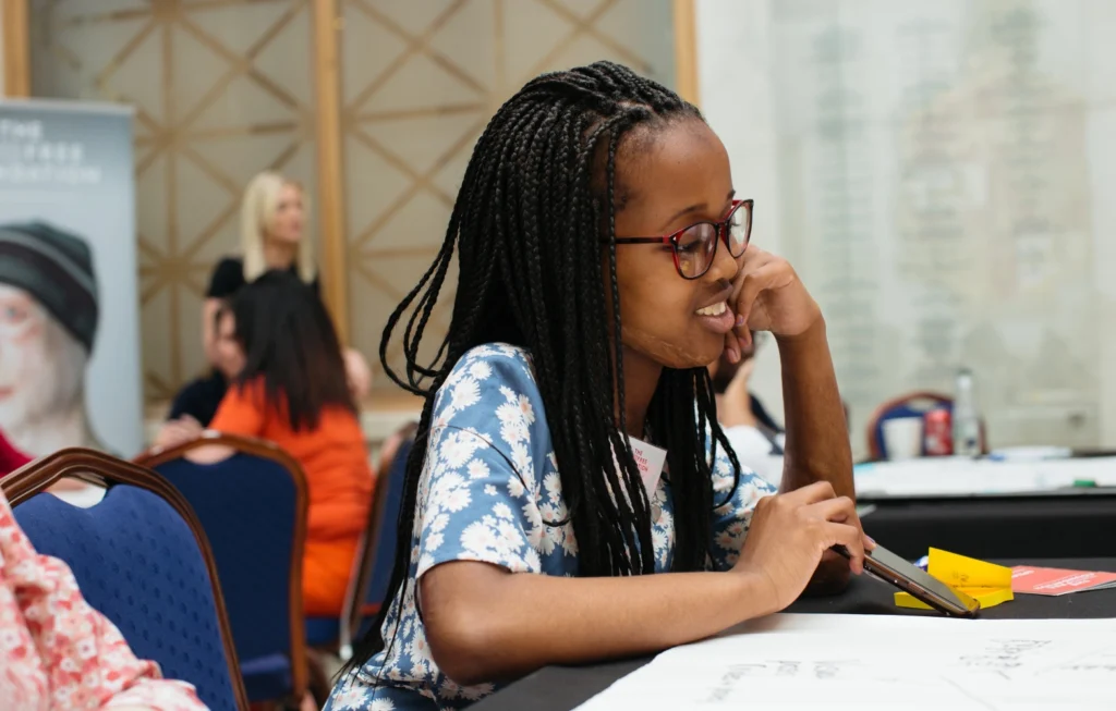 Young woman with braids and glasses smiling while seated at a table during an event or workshop.