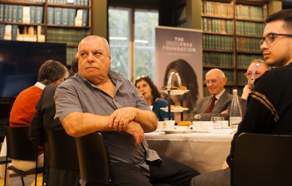 Man in blue shirt sitting at panel event in library setting with other participants and audience members visible.