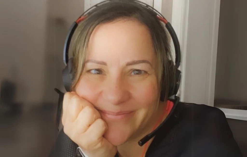 Woman wearing gaming headset with red accents, smiling at camera while resting chin on hand.