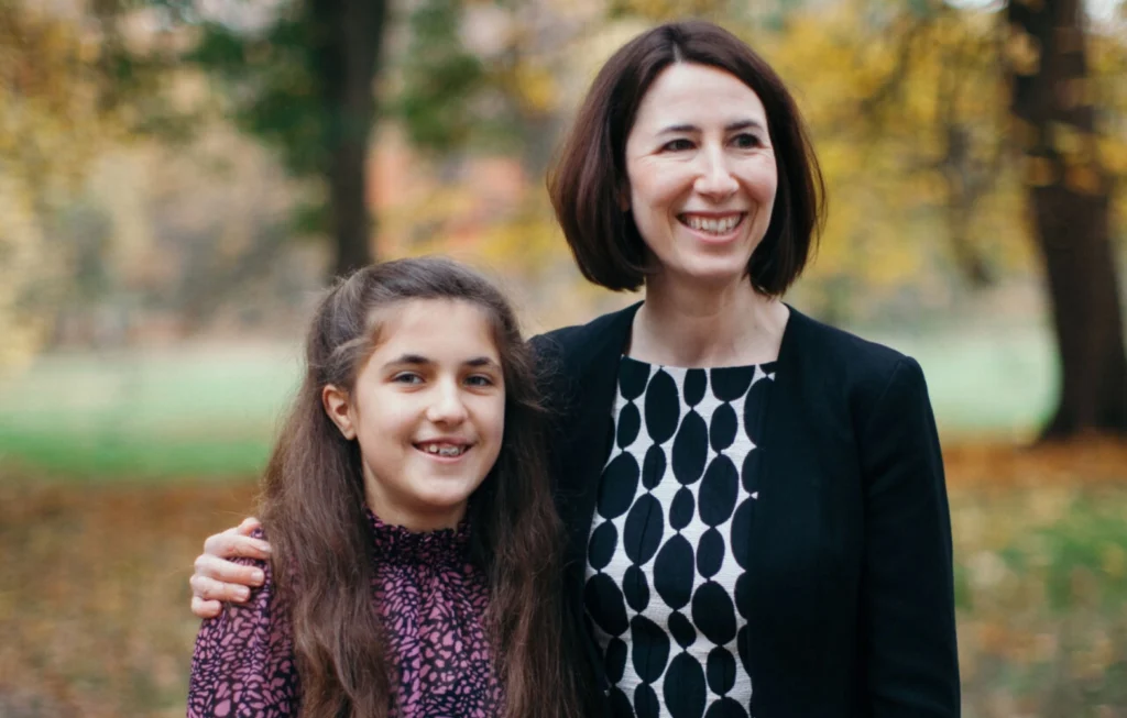 Woman and young girl smiling together outdoors in autumn setting with trees in background.