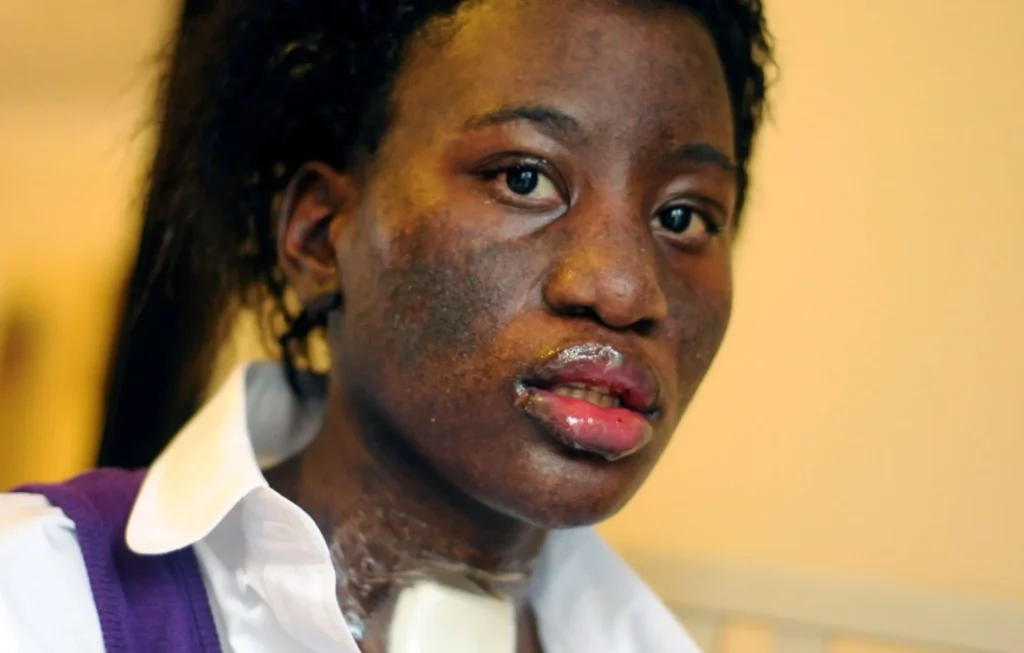 Woman wearing white and purple shirt against yellow background, looking toward camera.