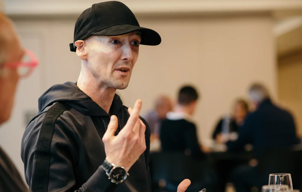 Man in black cap and jacket gesturing while speaking at an indoor event with people in background.