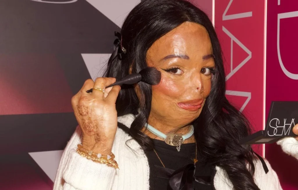 Woman applying makeup with brush, wearing white jacket and gold jewellery at event with pink backdrop.