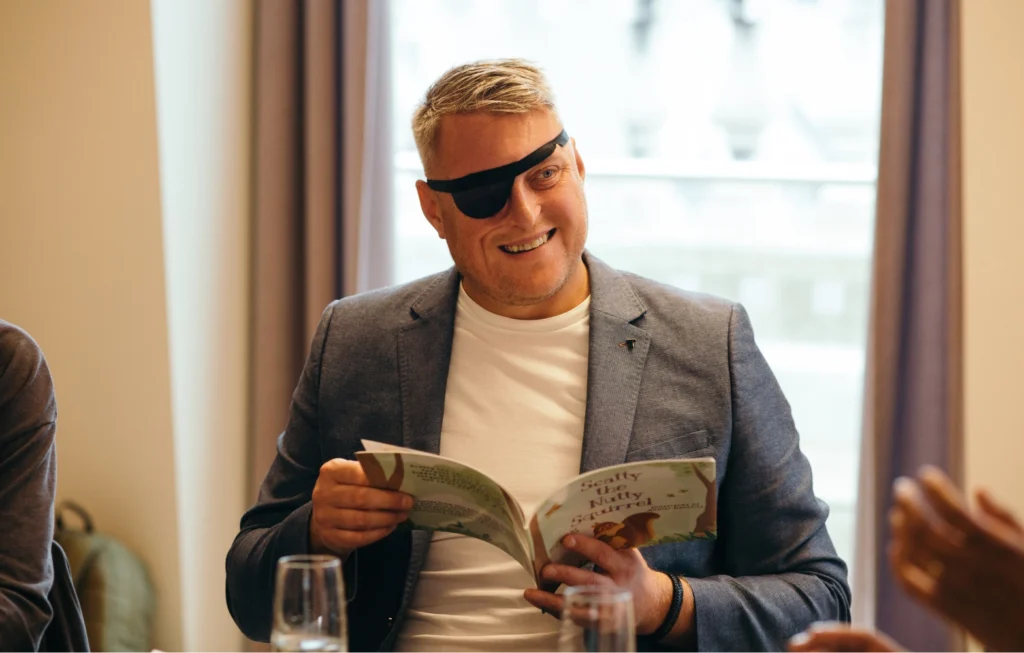 Man wearing eye patch and blazer reading a book at an indoor event with large windows behind him.