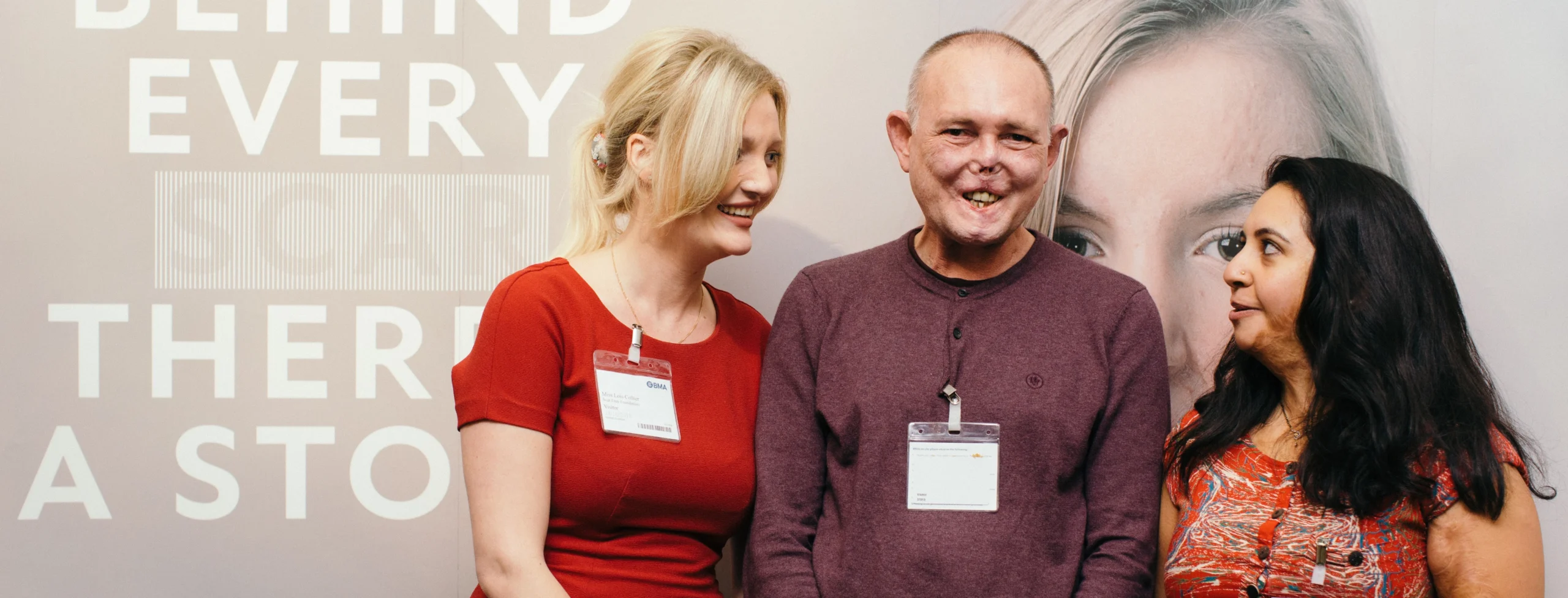 Three people wearing name badges stand smiling in front of a wall with text about stories behind scars.