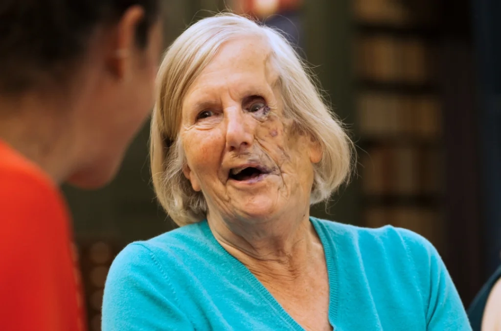 Older woman with white hair wearing a turquoise top, speaking and smiling during a conversation.