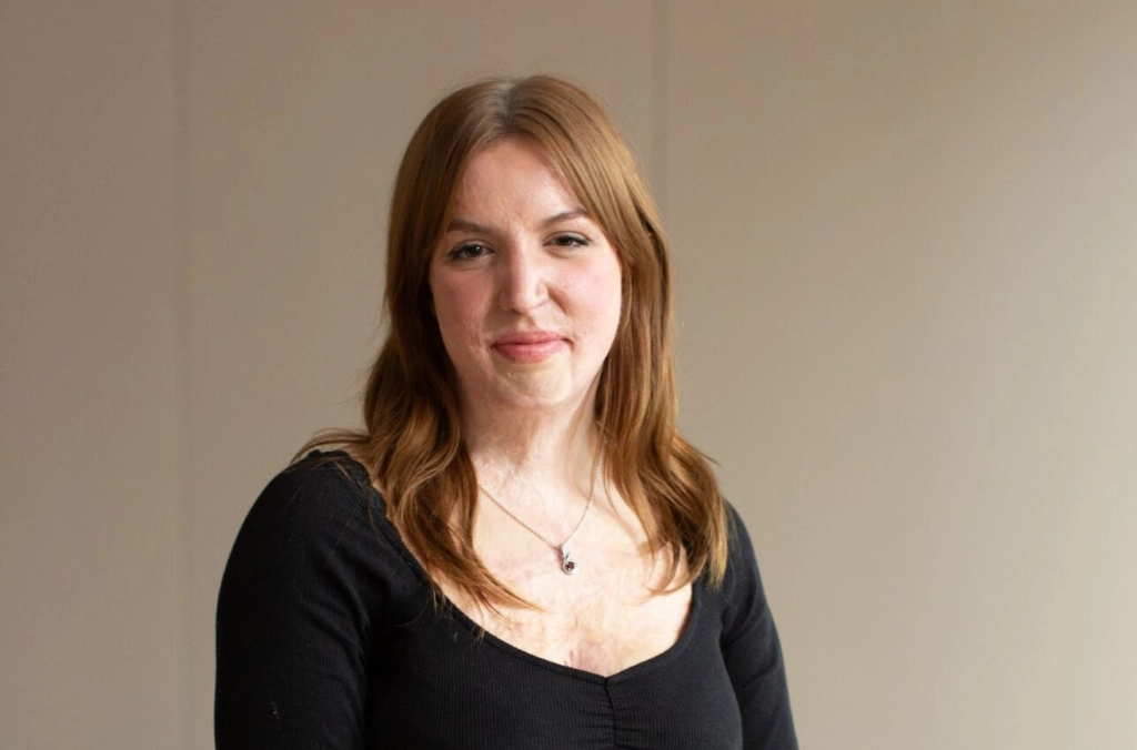 Woman with reddish-brown hair wearing a black v-neck top, smiling at camera against neutral background.