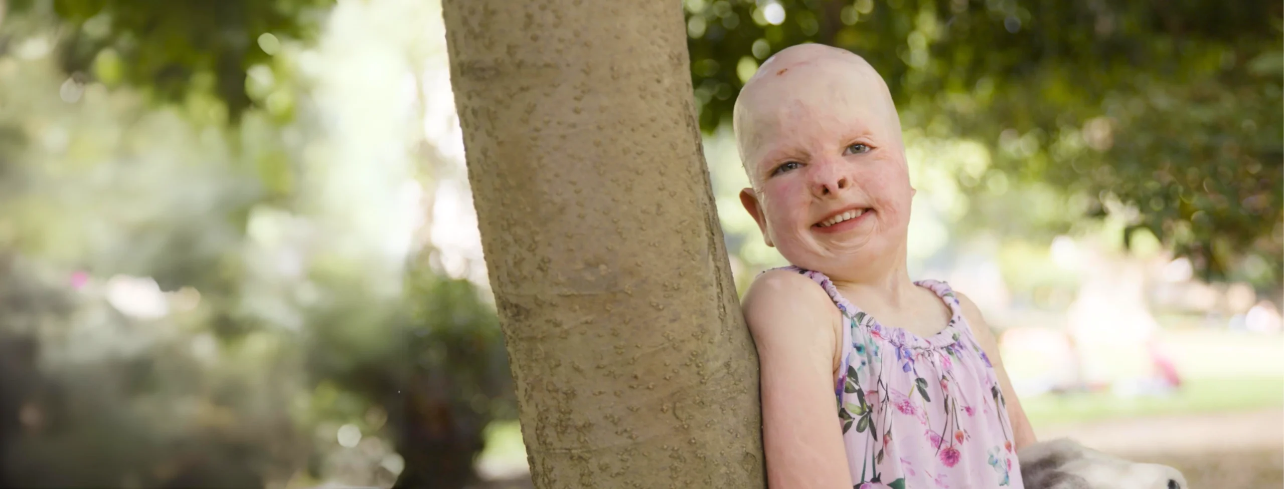 Young child standing by a tree, smiling at the camera in outdoor setting.