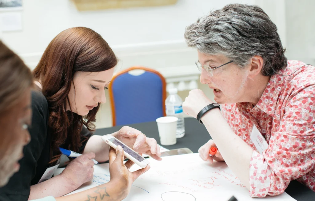 A woman in a pink floral shirt discusses medical research with a younger woman holding a tablet at a table.
