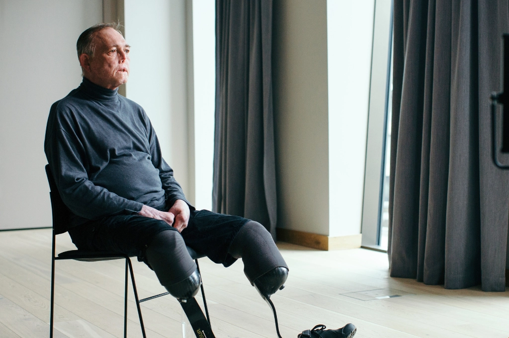 Man with prosthetic leg sitting on chair in bright room, looking toward window thoughtfully.